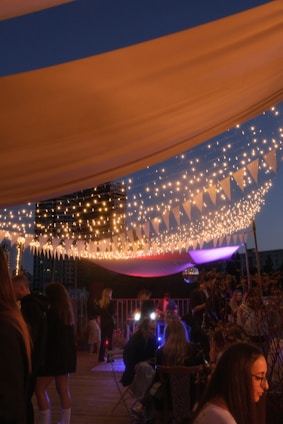 A nighttime outdoor party with string lights forming a canopy, creating a warm and festive atmosphere. People are mingling and sitting in groups under the decorative lights, with a backdrop of city buildings. A mix of colorful soft lights adds vibrancy to the scene.