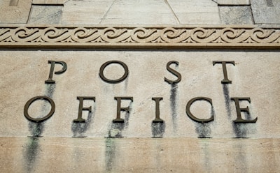 Wall inscription with large, bold letters spelling out 'Post Office', set against a textured stone surface with decorative carvings above.