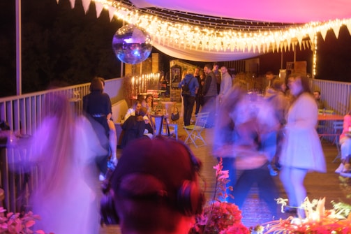 A lively outdoor party scene with people socializing and dancing under warm string lights. A disco ball hangs overhead, reflecting light across the area. Several tables and chairs are arranged along the space, some occupied by seated guests. The atmosphere has a colorful and vibrant ambiance.