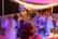 A lively outdoor party scene with people socializing and dancing under warm string lights. A disco ball hangs overhead, reflecting light across the area. Several tables and chairs are arranged along the space, some occupied by seated guests. The atmosphere has a colorful and vibrant ambiance.