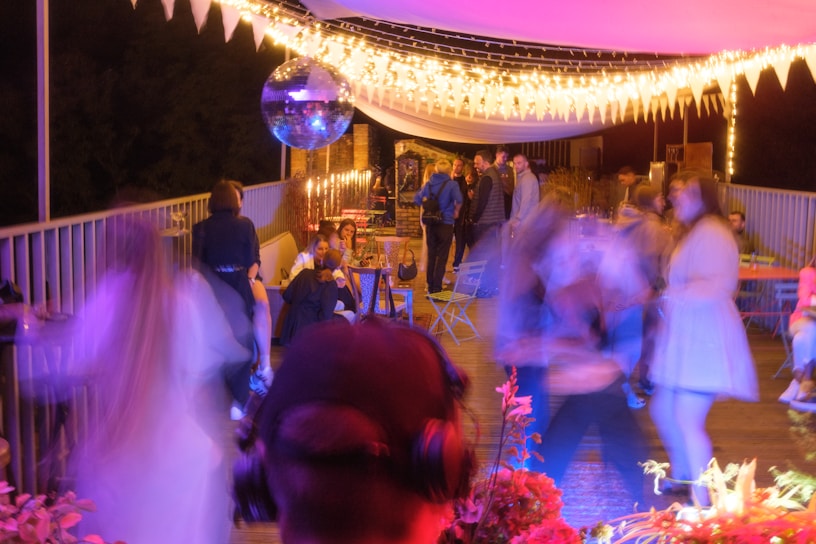 A lively outdoor party scene with people socializing and dancing under warm string lights. A disco ball hangs overhead, reflecting light across the area. Several tables and chairs are arranged along the space, some occupied by seated guests. The atmosphere has a colorful and vibrant ambiance.