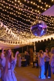 A lively outdoor party scene with several people socializing and dancing under string lights. A prominent disco ball hangs from above, adding a festive atmosphere. The area is decorated with white triangular pennants and soft lighting creates a warm ambience.