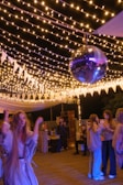 A lively outdoor party scene with several people socializing and dancing under string lights. A prominent disco ball hangs from above, adding a festive atmosphere. The area is decorated with white triangular pennants and soft lighting creates a warm ambience.