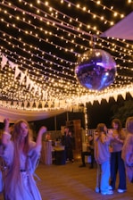 A lively outdoor party scene with several people socializing and dancing under string lights. A prominent disco ball hangs from above, adding a festive atmosphere. The area is decorated with white triangular pennants and soft lighting creates a warm ambience.