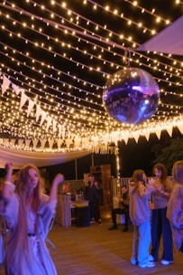 A lively outdoor party scene with several people socializing and dancing under string lights. A prominent disco ball hangs from above, adding a festive atmosphere. The area is decorated with white triangular pennants and soft lighting creates a warm ambience.