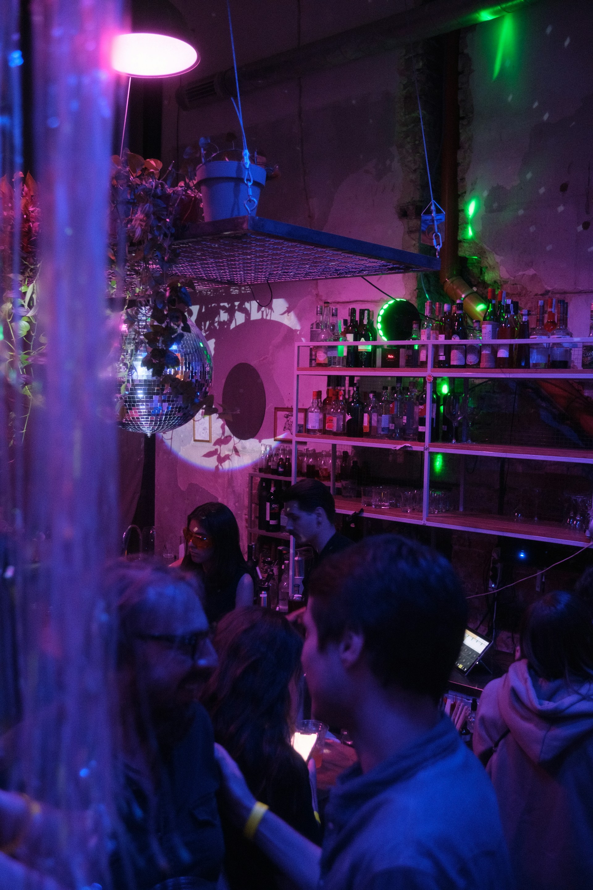 A dimly lit bar setting features several people socializing. The room is decorated with a disco ball, hanging plants, and shelves stocked with various bottles. Colored lights cast purple and green hues throughout the space, creating an ambient, vibrant atmosphere.
