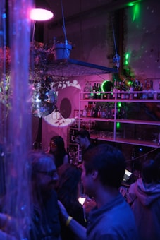 A dimly lit bar setting features several people socializing. The room is decorated with a disco ball, hanging plants, and shelves stocked with various bottles. Colored lights cast purple and green hues throughout the space, creating an ambient, vibrant atmosphere.