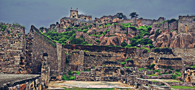 A historic fort sits atop a rocky hill, surrounded by ancient stone walls and ruins. The rugged landscape is dotted with green vegetation, contrasting with the weathered brown and gray stones. The fort features battlements and towers, providing a glimpse into its defensive architecture.