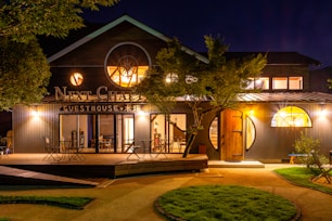 A spacious and welcoming guesthouse surrounded by lush greenery. The exterior is illuminated warmly against the night sky, featuring circular windows and a charming wooden deck with seating arrangements for guests.