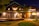A spacious and welcoming guesthouse surrounded by lush greenery. The exterior is illuminated warmly against the night sky, featuring circular windows and a charming wooden deck with seating arrangements for guests.