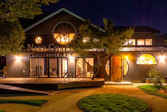 A spacious and welcoming guesthouse surrounded by lush greenery. The exterior is illuminated warmly against the night sky, featuring circular windows and a charming wooden deck with seating arrangements for guests.