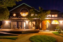 A spacious and welcoming guesthouse surrounded by lush greenery. The exterior is illuminated warmly against the night sky, featuring circular windows and a charming wooden deck with seating arrangements for guests.