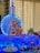 A colorful and vibrant setup featuring a statue of Lord Ganesha adorned with garlands, placed on a platform with blue and white decorative lights. A model rocket with 'ISRO' written on it forms the backdrop. Traditional lamps, candles, flowers, and fruits, including bananas and apples, are arranged around the deity.