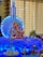 A colorful and vibrant setup featuring a statue of Lord Ganesha adorned with garlands, placed on a platform with blue and white decorative lights. A model rocket with 'ISRO' written on it forms the backdrop. Traditional lamps, candles, flowers, and fruits, including bananas and apples, are arranged around the deity.