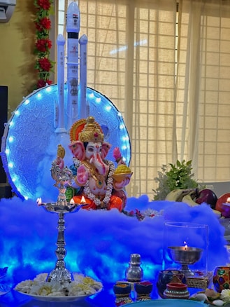 A colorful and vibrant setup featuring a statue of Lord Ganesha adorned with garlands, placed on a platform with blue and white decorative lights. A model rocket with 'ISRO' written on it forms the backdrop. Traditional lamps, candles, flowers, and fruits, including bananas and apples, are arranged around the deity.