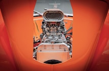 An up-close view of a polished, chrome car engine with intricate mechanics and details. The engine is framed by vibrant orange bodywork.