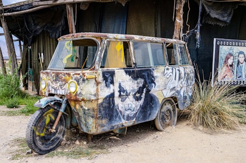 An old, rusty vehicle is creatively modified with artwork covering its surface. The vehicle's hood features a mischievous face with cartoonish eyes and a wide grin. A weathered and artistic design is seen along the side panels, and the vehicle appears to be parked in a rustic outdoor setting with dirt ground and grass. To the right, a poster with two people adds a contrasting element to the scene.