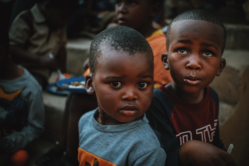 Two young children are sitting together, their faces displaying curiosity and seriousness. One child is wearing a grey shirt, and the other is in a shirt with text. The background includes other children and a plate of food.