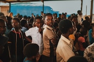 A group of children gather closely together in an outdoor setting, some looking directly at the camera while others are engaged in different activities. The environment appears lively and filled with a mixed group of kids wearing casual clothing. A blue tarp and wooden beams are visible in the background, suggesting a makeshift or temporary shelter.