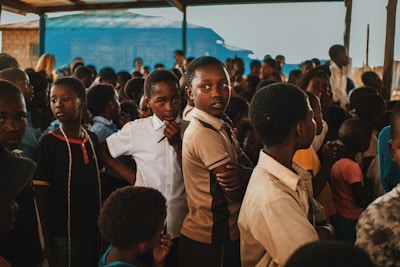 A group of children gather closely together in an outdoor setting, some looking directly at the camera while others are engaged in different activities. The environment appears lively and filled with a mixed group of kids wearing casual clothing. A blue tarp and wooden beams are visible in the background, suggesting a makeshift or temporary shelter.