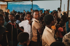 A group of children gather closely together in an outdoor setting, some looking directly at the camera while others are engaged in different activities. The environment appears lively and filled with a mixed group of kids wearing casual clothing. A blue tarp and wooden beams are visible in the background, suggesting a makeshift or temporary shelter.