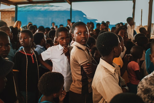 A group of children gather closely together in an outdoor setting, some looking directly at the camera while others are engaged in different activities. The environment appears lively and filled with a mixed group of kids wearing casual clothing. A blue tarp and wooden beams are visible in the background, suggesting a makeshift or temporary shelter.