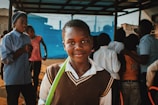 A young boy stands in the foreground with a slight smile, wearing a brown sweater vest over a white shirt. He holds a green object or ribbon. In the background, several other children and teenagers are present, engaged in various activities. They are under a shelter or roof, and a vivid blue background is visible.