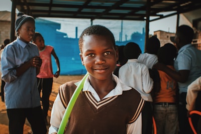 A young boy stands in the foreground with a slight smile, wearing a brown sweater vest over a white shirt. He holds a green object or ribbon. In the background, several other children and teenagers are present, engaged in various activities. They are under a shelter or roof, and a vivid blue background is visible.
