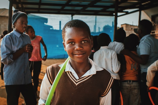 A young boy stands in the foreground with a slight smile, wearing a brown sweater vest over a white shirt. He holds a green object or ribbon. In the background, several other children and teenagers are present, engaged in various activities. They are under a shelter or roof, and a vivid blue background is visible.