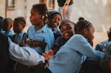 A group of children in blue uniforms sitting closely together, smiling and interacting in an outdoor setting. One child looks directly at the camera with a big smile, while others engage with each other. An adult's hand and legs are partially visible in the background.