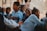A group of children in blue uniforms sitting closely together, smiling and interacting in an outdoor setting. One child looks directly at the camera with a big smile, while others engage with each other. An adult's hand and legs are partially visible in the background.