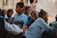 A group of children in blue uniforms sitting closely together, smiling and interacting in an outdoor setting. One child looks directly at the camera with a big smile, while others engage with each other. An adult's hand and legs are partially visible in the background.
