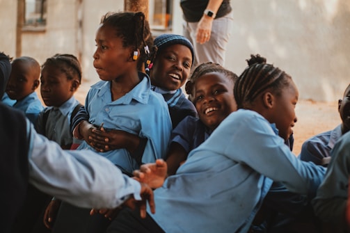 A group of children in blue uniforms sitting closely together, smiling and interacting in an outdoor setting. One child looks directly at the camera with a big smile, while others engage with each other. An adult's hand and legs are partially visible in the background.