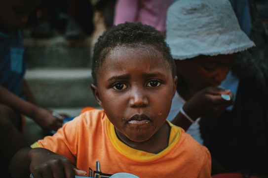 A young child with short hair and a serious expression stares directly at the camera. The child is wearing an orange shirt with a yellow collar. In the background, another person in white attire with a hat is seen eating with a spoon. The image appears to be taken outdoors with a group of people.