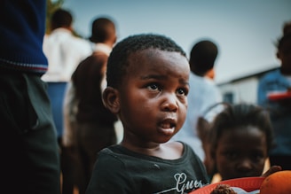 A young child with an expression of curiosity or concern holding a plate of food, surrounded by other children in an outdoor setting. The background is slightly blurred, emphasizing the child's face.