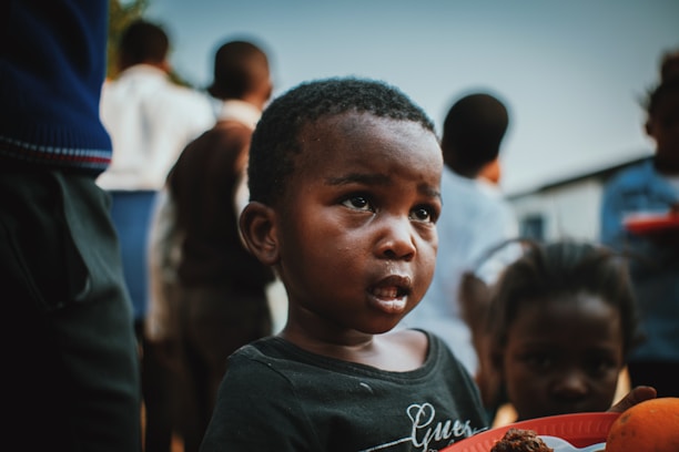 A young child with an expression of curiosity or concern holding a plate of food, surrounded by other children in an outdoor setting. The background is slightly blurred, emphasizing the child's face.