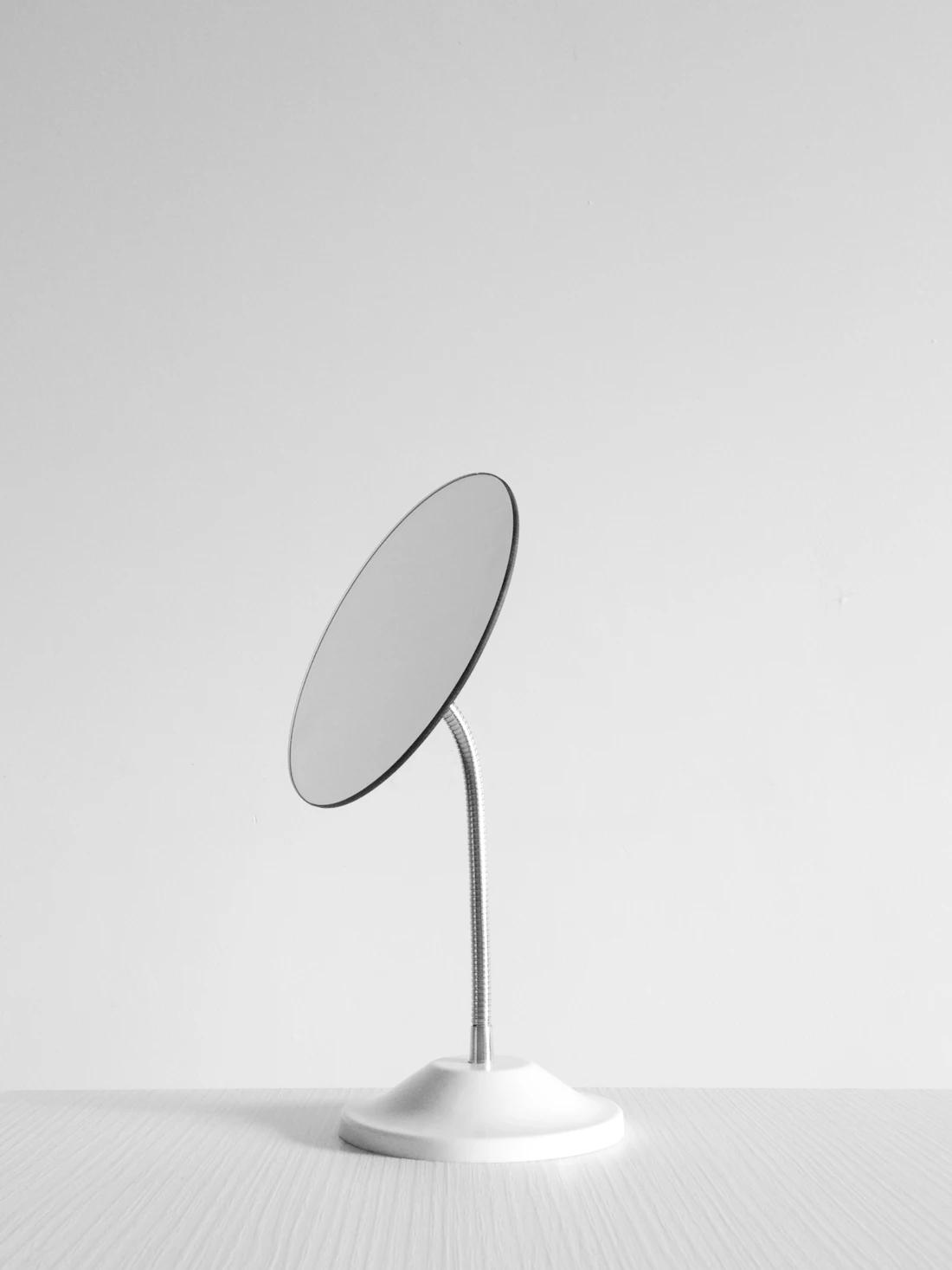 A small round mirror on a flexible stand against a plain white wall (grooming stand-in for upper-lip and chin hair removal)
