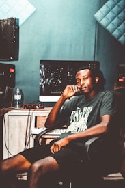 A person sits in a recording studio, surrounded by soundproof walls with acoustic panels. They are wearing a casual t-shirt and shorts. A computer monitor displays a dark screen with some visible reflections, and audio equipment is present, including speakers and a water bottle.