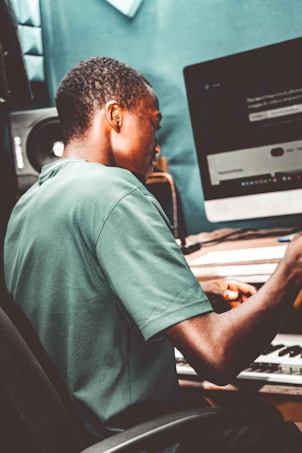 A person wearing a green shirt is seated at a desk, looking at a computer screen. Nearby is a keyboard and a speaker, suggesting a music production or creative workspace.