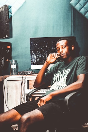 A young man sits in a recording studio, relaxed in a chair with his hand thoughtfully placed on his chin. He is wearing a casual t-shirt and shorts, and around him are various studio equipment including a computer screen, audio mixer, and a water bottle on the desk.