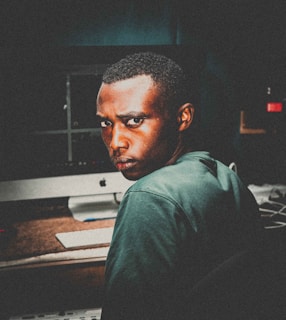 A person with a serious expression is sitting in front of a computer, looking towards the camera. The background is dimly lit, with the glow of the computer screen visible. The scene is indoors, with a moody and somewhat intense atmosphere.