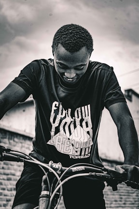 A person with a joyful expression is riding a bicycle, sporting a t-shirt that encourages positive thinking. The background includes a textured wall and a moody sky, adding contrast to the scene.