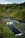 A serene outdoor swimming pool is nestled in a lush, green valley surrounded by rolling hills. The pool is adjacent to a small, white building with green doors. A gentle stream runs parallel to the pool, bordered by rocky terrain and vibrant moss-covered landscape.