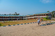 A two-story academic building has students and educators engaged on the grounds. The architecture is primarily brickwork with a blue roof. Several figures are grouped in a discussion near the architecture block. A paved walkway with blue and white edges leads towards the building, surrounded by a lawn with scattered trees and shrubs.