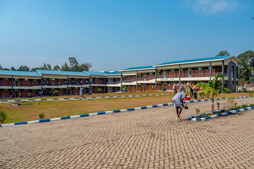 A two-story academic building has students and educators engaged on the grounds. The architecture is primarily brickwork with a blue roof. Several figures are grouped in a discussion near the architecture block. A paved walkway with blue and white edges leads towards the building, surrounded by a lawn with scattered trees and shrubs.