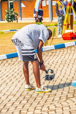 A person in casual attire is bending down to adjust or inspect a small object that appears to be a drone or camera equipment on a paved area. In the background, there are people wearing reflective vests and some survey equipment. The setting appears to be a sunny outdoor location with a combination of paved and grassy areas.