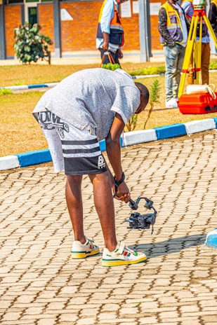 A person in casual attire is bending down to adjust or inspect a small object that appears to be a drone or camera equipment on a paved area. In the background, there are people wearing reflective vests and some survey equipment. The setting appears to be a sunny outdoor location with a combination of paved and grassy areas.
