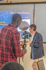 A person in a red plaid shirt operates a camera on a stabilizer, recording another individual who is speaking and gesturing. The background features a presentation slide with text that includes the word 'Ubiquitousness.' The speaker is wearing a dark blazer and light-colored pants.