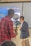 A person in a red plaid shirt operates a camera on a stabilizer, recording another individual who is speaking and gesturing. The background features a presentation slide with text that includes the word 'Ubiquitousness.' The speaker is wearing a dark blazer and light-colored pants.