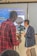 A person in a red plaid shirt operates a camera on a stabilizer, recording another individual who is speaking and gesturing. The background features a presentation slide with text that includes the word 'Ubiquitousness.' The speaker is wearing a dark blazer and light-colored pants.